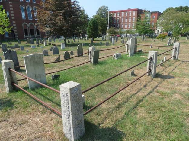 Fenced in plot in Eastern Cemetery with 6 granite corner and center posts and 2 iron posts connecting each on a sunny day. 2 red brick buildings frame the background behind green leafy trees and grass and many scattered old slate and marble grave markers