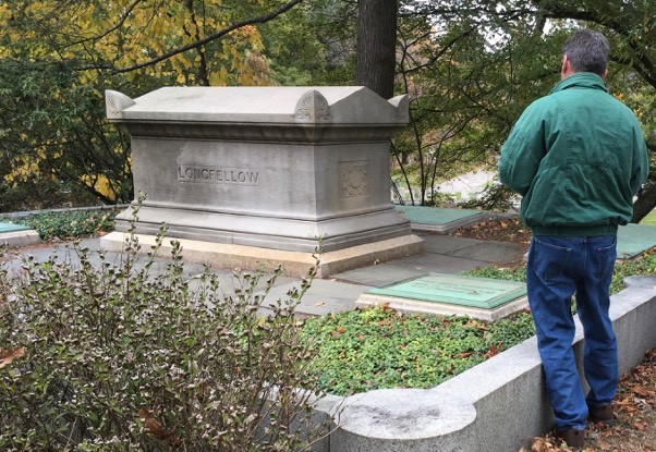 a person stands in front of a cemetery granite box monument with curbs within a leafy and grassy area