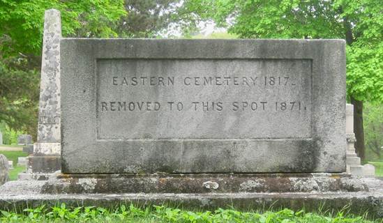 large granite box grave marker set amid the backdrop of leafy green trees, a tall weathered marble column grave marker, and green grass, inscribed Eastern Cemetery 1817, Removed to this spot 1871.