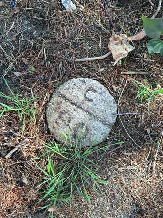 A round grey concrete marker on the ground amid grass and dirt. A weathered carving: C above and 90 below a bisecting line.