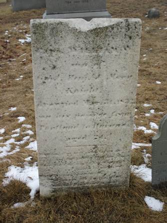 Chipped, weathered, and eroded rectangular marble marker sprinkled with black lichen. With carving hard to make out, the marker is set among other slate grave markers in the winter ground where brown grass shows through the patches of snowfall.