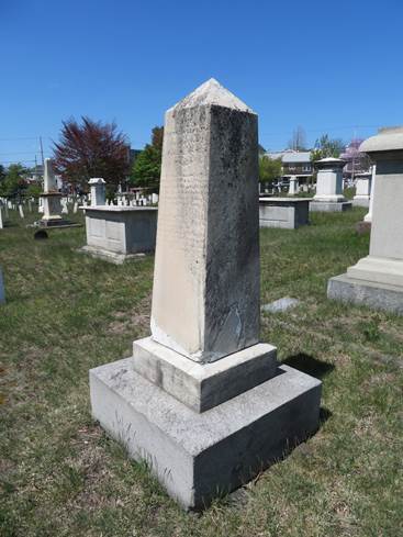 Weathered and inscribed marble obelisk grave monument, dirtied with black organic matter, set on 2 granite plinths among other large monuments in a grassy area.