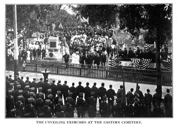 The unveiling Exercises at the Eastern Cemetery for the Alonso Stinson monument. A large crowd of uniformed men faces a large crowd behind a fence that has gathered in the cemetery around the large monument.