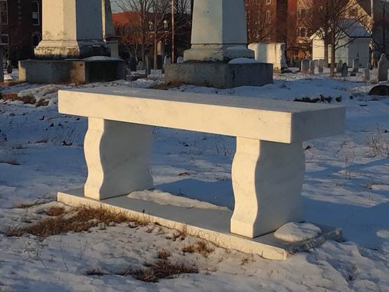 Marble bench set among large monuments and a snowy ground. The Dead House of Eastern Cemetery is in the background.