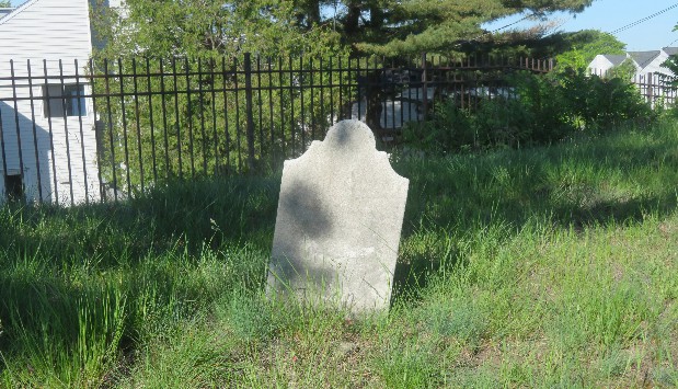 old marble gravestone in grassy green a cemetery with a black metal fence and green leafy trees behind it