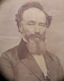 Old photograph of Joseph Thompson who looks into the camera, a white man with tousled short hair combed up and back with a neck-length bushy beard and mustache. He wears a top coat with wide lapels and a white shirt under it.