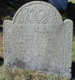 old slate curved gravestone carved with a winged skull, decoration on the sides, and an inscription