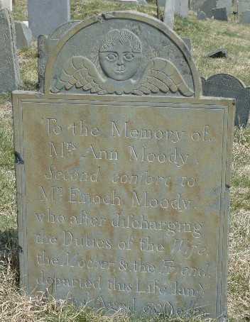 old slate gravestone among other stones in a grave yard carved with a winged face and an inscription