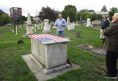 a person stands behind a box tomb draped with a US flag in Eastern Cemetery while others look on