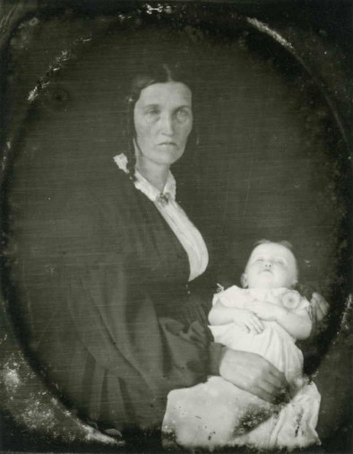 portrait of a frowning Hannah Strout with a long dark hair ringlet resting on her frilly white dress collar, and a dark coat over it who holds a baby wearing a long white dress whose arms are crossed and eyes are closed on her lap