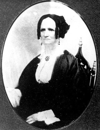 portrait of a frowning Dolly Pease sitting in a chair with 2 finials on the tips of its back who is wearing long earrings, a jeweled pin in a flower shape on her neck and a high-necked white frilly top under a dark coat