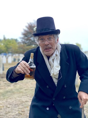 Billy Hans wears a top hat, double breasted coat, and wire-framed glasses down his nose gestures with a bottle of brown liquid and corked top his hand to come closer to him
